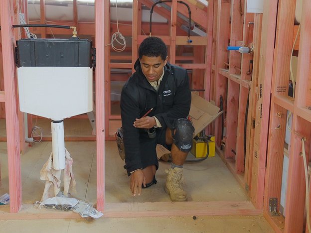 A young man kneels on the floor of a wooden building frame. He holds a pencil in his hand and has a tool belt around his waist.