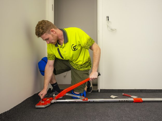 A male kneels on carpet and presses it onto the floor.