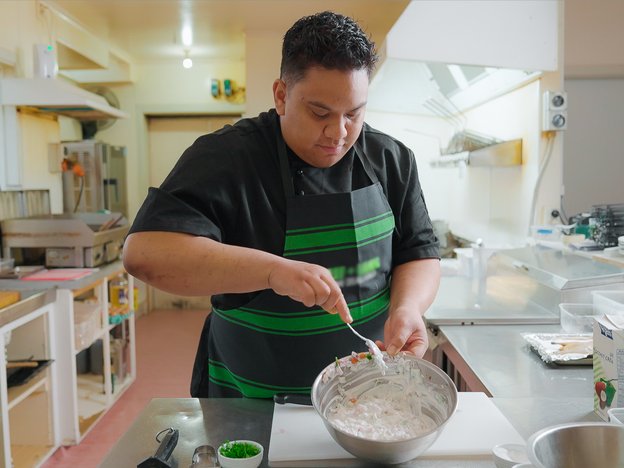 A chef combines ingredient in a mixing bowl. Utensils and ingredients can be seen on the benchtop.