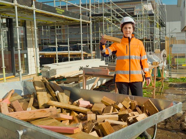 A woman in an orange hi-vis and safety helmet holds a piece of wood. In front of her is a container full of wood.