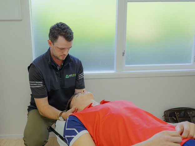 Man in navy vest and green pants massages a patient's neck. The patient lies on his back on a chiropractic table.