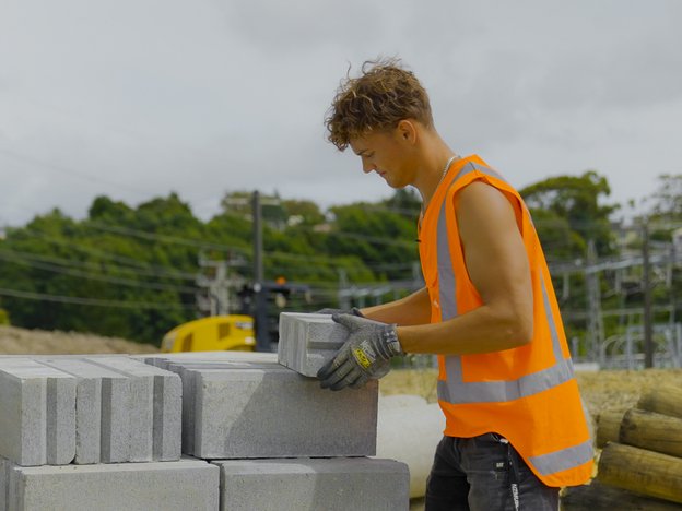 A young man stacks bricks at a construction site. He wears an orange vest and grey gloves.