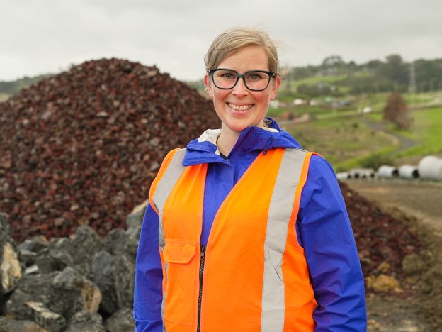 A person stands in front of rock piles. They wear an orange hi-vis.