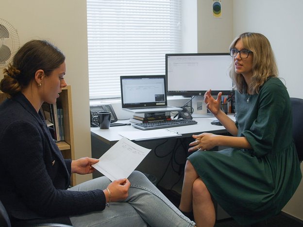 A woman interacts with a young female client in an office. The young woman holds a piece of paper.