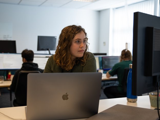 A computer engineer sits at an office desk looking at a monitor. Behind them are two colleagues facing the opposite direction, doing the same.
