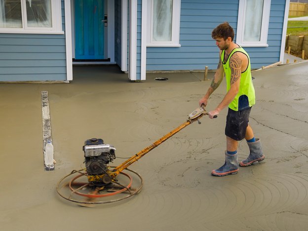 A man in a hi-vis uses a powered concrete float to smooth cement in front of a blue house.