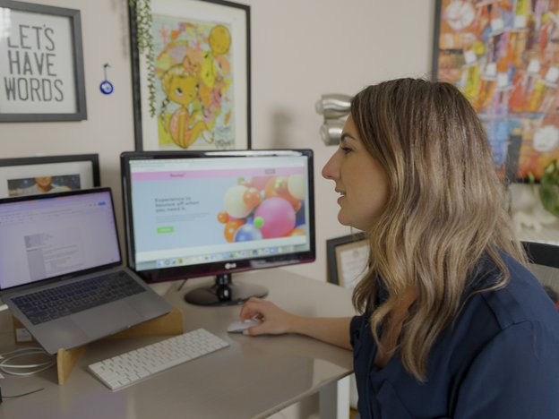 A woman sits at a desk using a laptop and mouse. There is a computer monitor on the table next to her. She is in a home office.