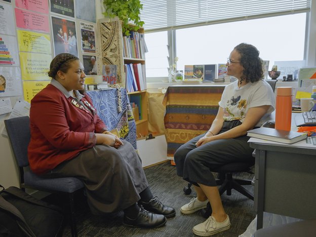 A female wears a school uniform and talks to another female who sits behind a desk.