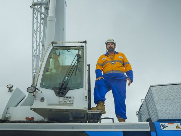 A man wears a blue and orange suit with work boots and a white helmet. He stands on top of a crane.