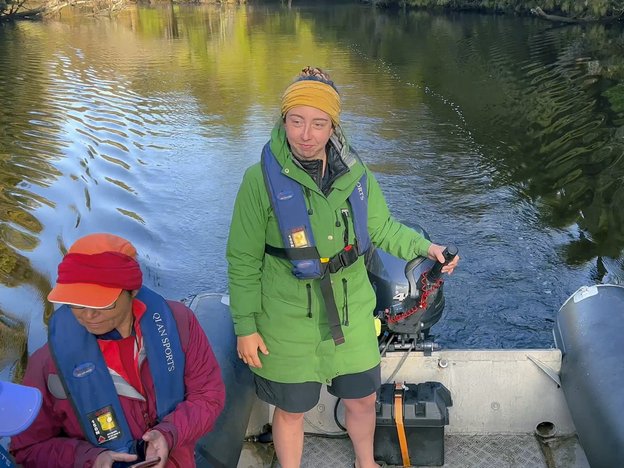 A female deckhand stands in a boat on a river. Two women sit down beside her. They all wear life jackets.