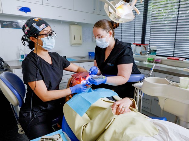 A dentist and dental assistant inspect the mouth of patient using tools. The dentist wears a white lab coat and the dental assistant wears blue scrubs. They both wear hair nets and white gloves.