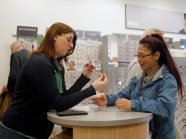 A female dispensing optician assists a woman with her glasses. They are in an optometrist store.