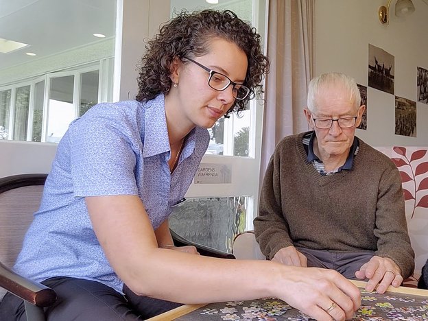 A female picks up a puzzle board piece and sits next to an elderly male.