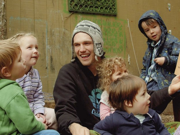 A male teacher sits with five young children. They are playing and laughing.