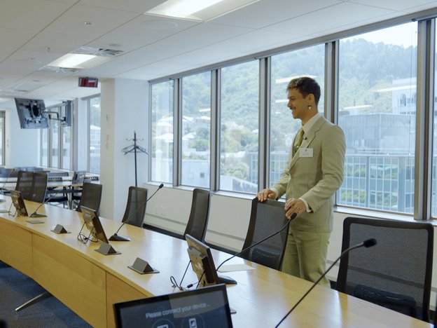 A man wears a suit and smiles as he holds a chair and looks at a desk.