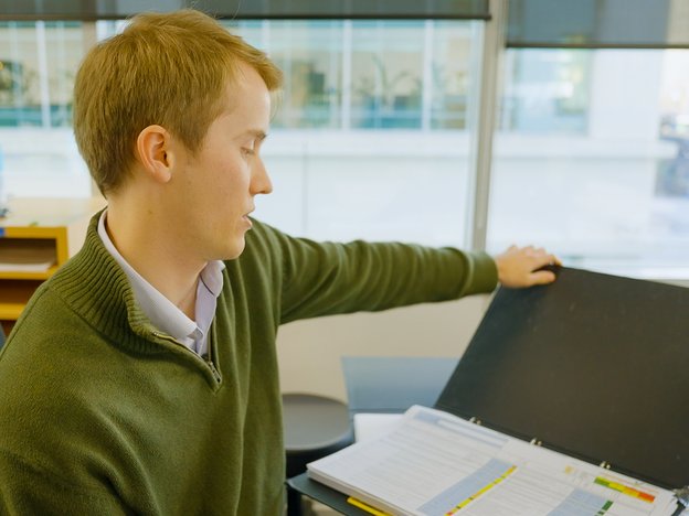 A young man sits in office reading a folder. He is wearing a green jersey.