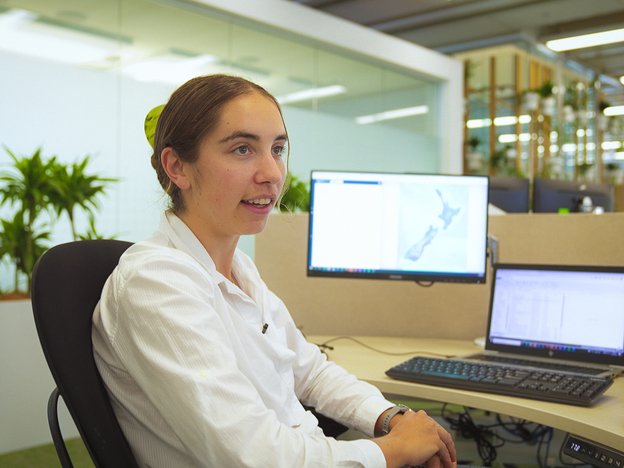 A young woman speaks at her office desk. A picture of New Zealand can be seen on one of her monitors.