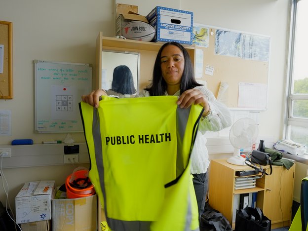 A woman stands in an office holding up a fluorescent yellow vest. The vest has the words 'public health' on the back.