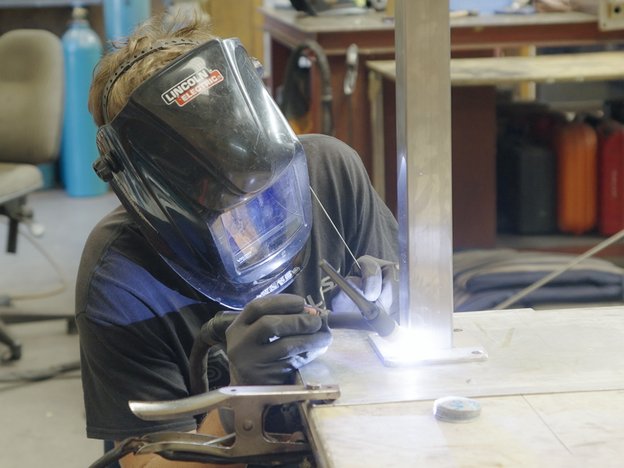 A man welds one piece of metal to another and wears gloves and eye protection.