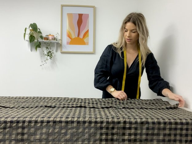 Woman with blonde hair in a black dress with measuring tape around her neck measures out checkered brown and black material on a table.