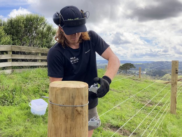 A man wears a navy polo shirt, navy cap, gloves and earmuffs. He fixes a wire fence to a wooden pole in a grass paddock.