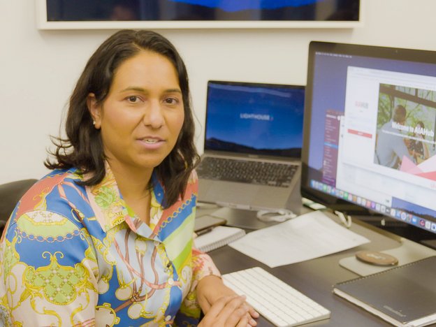 A woman looks into the camera while sitting at a desk. A laptop, computer monitor, keyboard and notepad are on the desk in front of her.