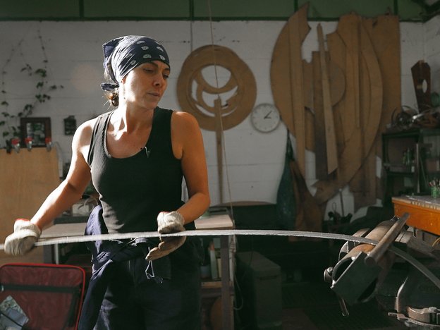 A woman holds a long, thin strip of metal in an art workshop. She is wearing white gloves and a navy-blue bandana.