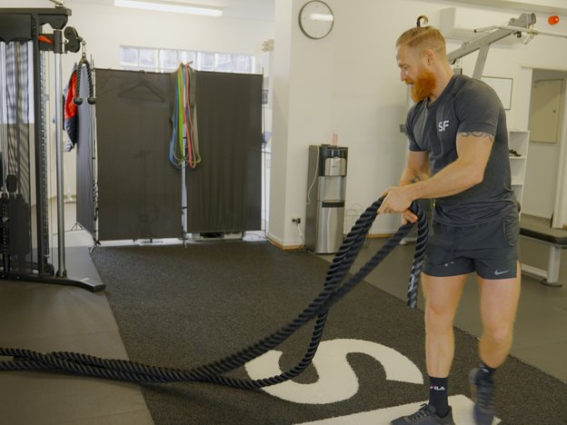 Male in navy t-shirt and shorts and sneakers with a ginger beard uses ropes to work out on a mat in a gym. He faces side on to the camera.