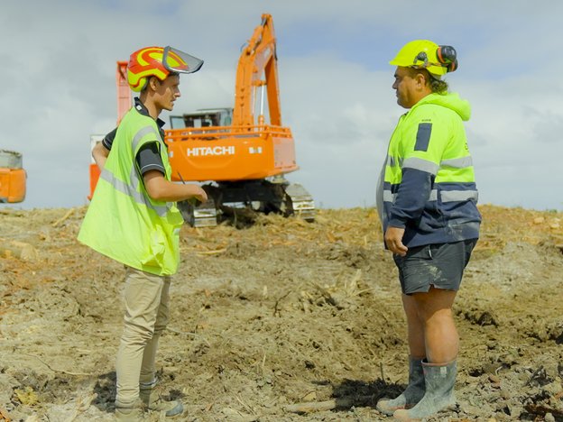 Two males in fluorescent vests and helmets stand at a construction site. An orange crane is in the background.