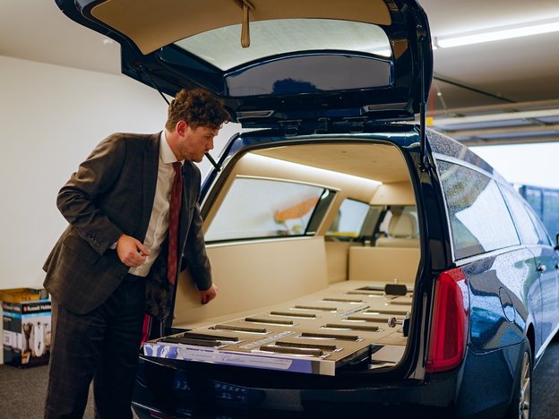 A man in a suit looks into the open boot of a hearse.