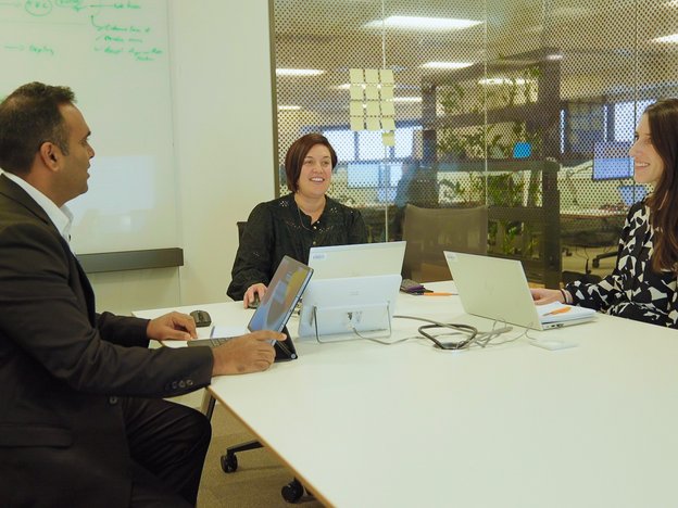 A man and two women sit around a table interacting in a meeting room. They have laptops in front of them.
