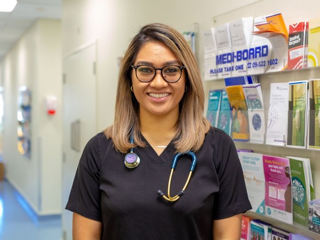 A smiling female doctor stands in the hallway of a hospital. She wears a stethoscope around her neck.
