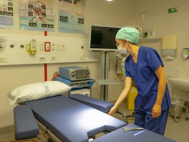 A female surgeon prepares abed in a hospital room. She wears a face mask and green hairnet. There are surgical scissors on the bed.