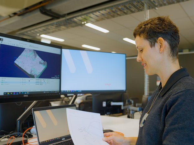 A woman sits at a desk in front of two computer monitors and a laptop. She is holding a document and reviewing it.