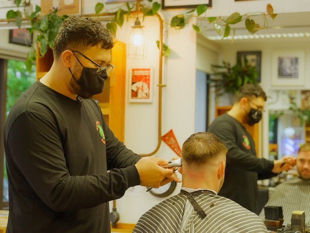 A male barber wearing a face mask shaves a man's head while standing in a salon. A mirror shows their reflection behind them.