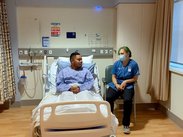 A male sits in a hospital bed and talks to a female.