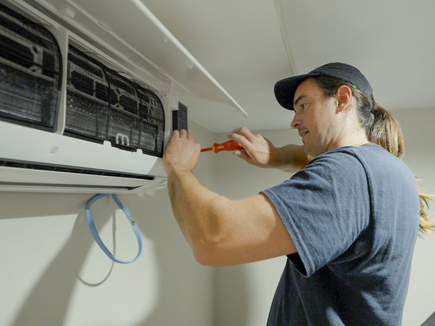 A young man repairs a wall-mounted heat pump using a screw driver.
