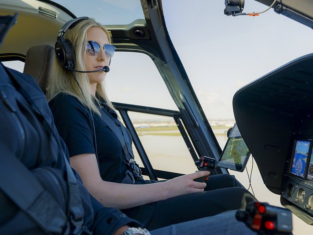 A female pilot sits in the cock pit of a helicopter. She wears a headset and holds a gear stick.
