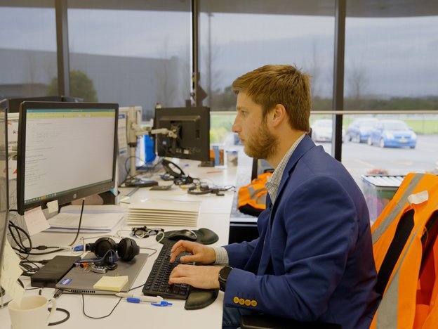 A man sits at a desk looking at two computer monitors. He is typing on a keyboard and using a mouse. He wears a suit, and an orange vest hangs on the back of his chair.