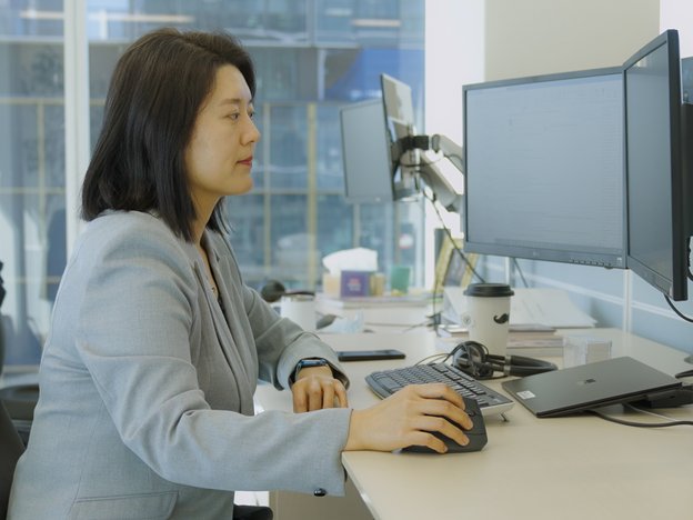 A person sits at a desk using a computer that has two monitors. They are in a modern office space near the window.