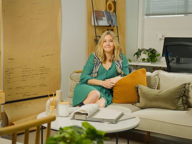 A young woman sits on a couch in a trendy work space. Behind her is an office desk and a large brown sheet of paper hangs from the wall.