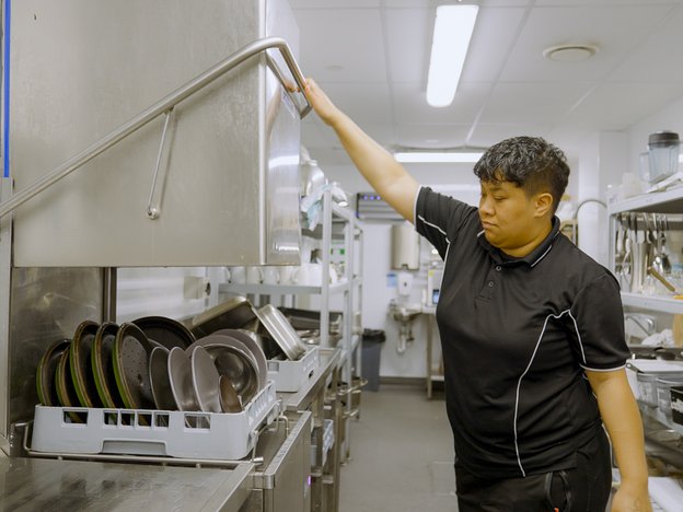 A woman uses a dishwasher to clean a tray of plates in a commercial kitchen.