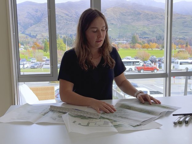 An architect looks at plans at a work desk. They are sitting in a room wearing a black shirt.