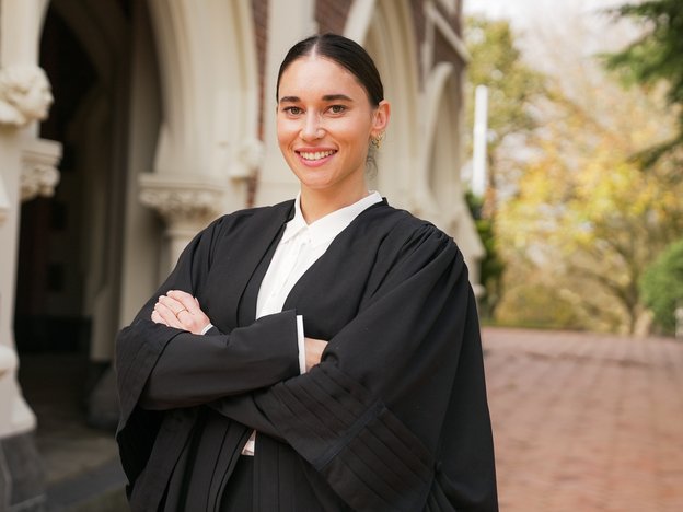 A woman stands outdoors with folded arms in front of a building. She wears a black legal gown.