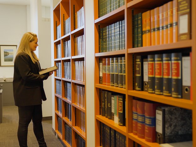 A woman stands side-on holding a book in a legal library. She is facing a large wall of books.