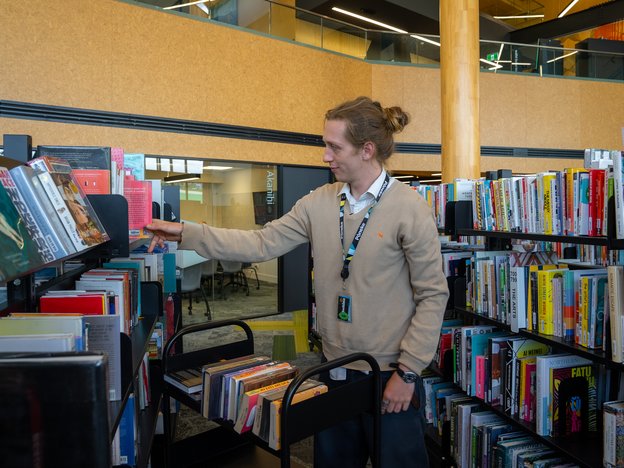 A librarian places books back into shelves. They are wearing a brown sweater and black pants.