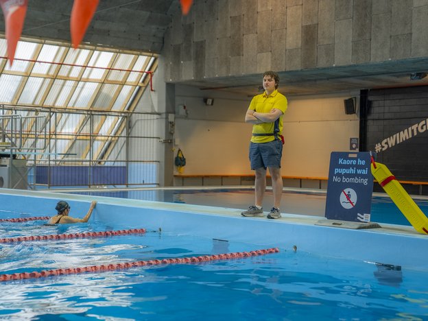 A male lifeguard starts at the head of a swimming pool with folded arms in a swimming venue.