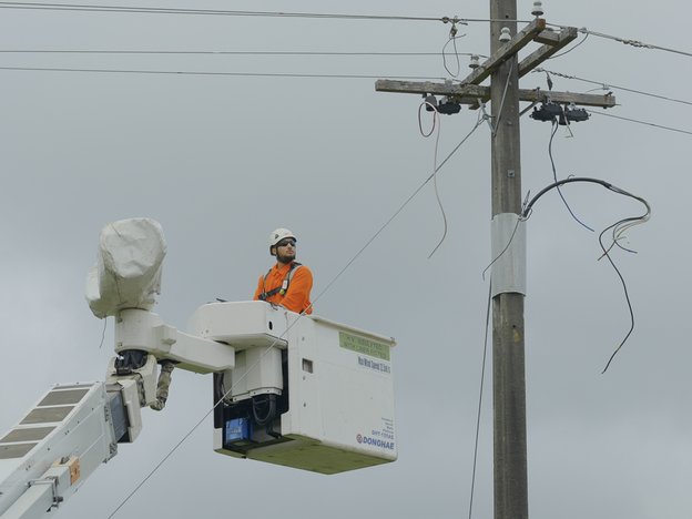 A man stands in the bucket of a mobile elevated work platform. He is under some power lines. He wears a white helmet and orange uniform.