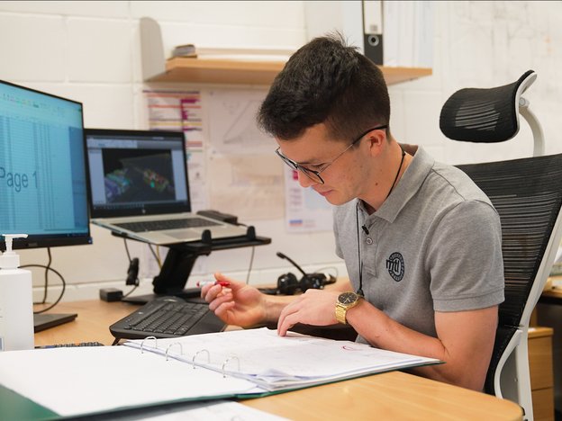 A man sits and looks at a document in a folder on a desk. He holds a pen in one hand and there is an open laptop and monitor on the desk.