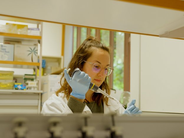 A woman uses a device to transfer a solution to a test tube. She is wearing a lab coat and blue gloves.